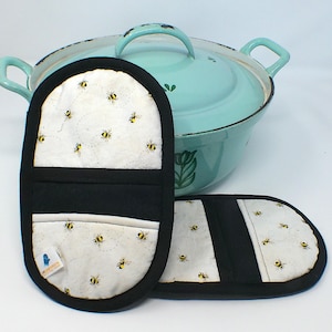 May include: Two black and white oven mitts with a bee pattern, next to a light blue pot with handles. The mitts have a black border and a white fabric with yellow and black bees. The pot has a lid and two handles.
