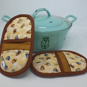 May include: A light blue Dutch oven with a lid, and two matching oven mitts. The mitts have a tan background with a pattern of teacups and brown trim. The oven mitts are next to the Dutch oven.