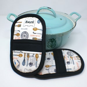 May include: Two black and white pot holders with a kitchen theme. The pot holders have a white background with black text and images of kitchen utensils. The text on the pot holders includes "Bake", "Gourmet Method", "Market Fresh", "Salad", and "Blend".