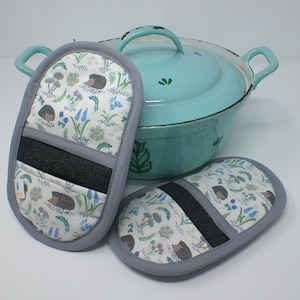 Puede incluir: Una olla de esmalte azul claro con tapa y dos manoplas a juego. Las manoplas tienen un fondo blanco con un estampado de erizos, setas y flores. Las manoplas tienen un borde gris y una banda negra.