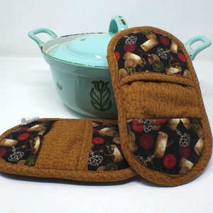 Könnte beinhalten: Zwei braune Topflappen mit einem schwarzen und roten gemusterten Stoff, der ein Traubenmuster zeigt. Die Topflappen haben eine Tasche auf der Rückseite.