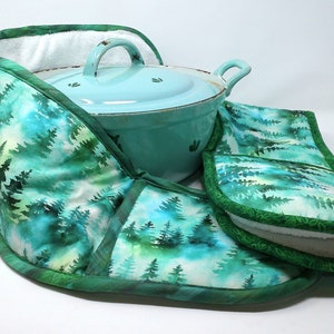 Puede incluir: Un juego de agarraderas de olla verdes y azules con un estampado de bosque de acuarela. Las agarraderas de olla están forradas con vellón blanco y tienen un ribete verde. Las agarraderas de olla se muestran con una olla azul claro.