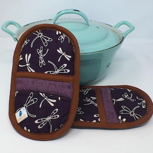 Könnte beinhalten: Zwei lila-braune Topflappen mit Libellenmuster, neben einem hellblauen Topf. Die Topflappen haben einen braunen Rand und einen lila Akzent. Das Libellenmuster ist weiß.