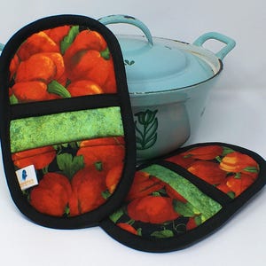 以下が含まれることがあります： 黒い背景に赤いカボチャの模様が施された2つのオーブンミトンと鍋つかみ。緑と黒の縁取りが施されています。背景には、取っ手が付いた水色の鍋があります。