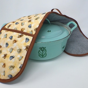 Puede incluir: Una olla holandesa azul claro con un guante de horno a juego. El guante de horno tiene un fondo amarillo con un patrón de tazas de café y ribetes marrones. La olla holandesa tiene un diseño floral verde en el lateral.