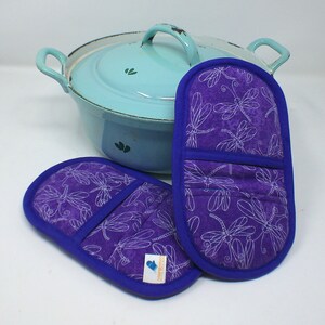 Könnte beinhalten: Zwei violette Topflappen mit einem weißen Libellenmuster und blauem Besatz. Die Topflappen liegen auf einer weißen Oberfläche neben einem hellblauen Emailletopf mit einem weißen Libellenmuster.