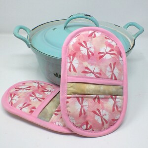 Könnte beinhalten: Zwei rosa Topflappen mit einem Libellenmuster. Die Topflappen haben einen hellrosa Hintergrund mit weißen Libellen und einem rosa Rand. Die Topflappen liegen vor einem hellblauen Topf mit Deckel.