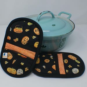 以下が含まれることがあります： ハロウィーンのかぼちゃ模様の黒いオーブンミトンと鍋つかみ。ミトンは、オレンジ色のかぼちゃと葉っぱの黒い背景です。背景には、蓋付きの薄い青色の鍋があります。