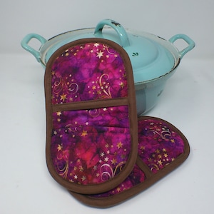 May include: A pair of purple and gold pot holders with a brown trim. The pot holders have a star and swirl pattern.