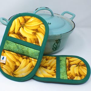 Könnte beinhalten: Zwei grüne und gelbe Topflappen mit Bananenmotiv und einem hellblauen Topf im Hintergrund. Die Topflappen haben einen Bananendruck mit einem grünen Akzentstreifen. Der Topf hat eine hellblaue Farbe mit einem grünen Blattmuster.