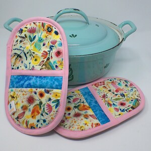 Könnte beinhalten: Zwei gesteppte Ofenhandschuhe mit floralem Muster und einem hellblauen Streifen, rosa eingefasst, vor einem hellblauen Topf mit Deckel und Griffen. Die Handschuhe haben ein farbenfrohes Design mit Blumen, Vögeln und einem Hasen.