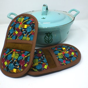 Könnte beinhalten: Zwei braune Topflappen mit einem bunten Muster aus Wollknäueln und Nadeln. Die Topflappen liegen auf einer weißen Oberfläche neben einem hellblauen Emailletopf mit grünem Blumendesign.