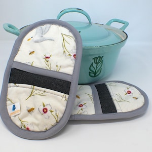 May include: Oven mitts with a floral and bee design, and a light blue Dutch oven. The mitts have a grey trim and a black heat-resistant band. The Dutch oven features a tulip design.