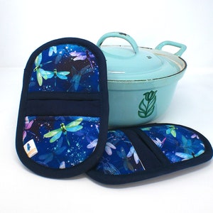 May include: Two blue and green dragonfly patterned oven mitts with a navy blue trim. The mitts are sitting on top of a light blue enamel Dutch oven.