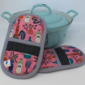 Könnte beinhalten: Rosa Ofenhandschuhe mit einem sich wiederholenden Muster aus Flaschen, Vasen und Pflanzen, mit grauem Rand und einem schwarzen hitzebeständigen Band. Ein hellblauer Kochtopf mit Deckel und Griffen befindet sich im Hintergrund.