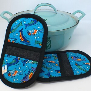 Puede incluir: Dos manoplas azules con borde negro y estampado de peces, junto a una olla azul claro. Las manoplas tienen un fondo azul con ilustraciones de peces coloridos en naranja, verde y morado. Una tira de tela negra está cosida en las manoplas.