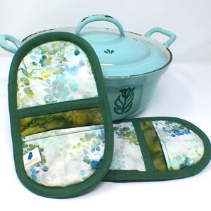 Può includere: Una pentola in smalto azzurro con coperchio e due presine abbinate. Le presine hanno un motivo floreale nei toni del blu, verde e bianco, con un bordo verde scuro. La pentola ha un disegno di tulipano sul lato.