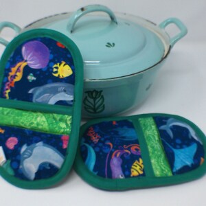May include: Two green pot holders with a blue background and a colorful underwater scene featuring seahorses, jellyfish, and dolphins. The pot holders are made of fabric and have a green border.