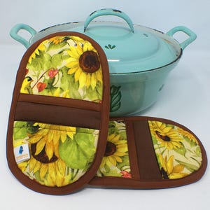 Könnte beinhalten: Ein hellblaues emailliertes Kochgeschirr mit Deckel und zwei passenden Ofenhandschuhen mit Sonnenblumenmuster. Die Handschuhe haben eine braune Umrandung und eine braune Tasche. Das Sonnenblumenmuster umfasst gelbe Sonnenblumen, grüne Blätter und rote Beeren.