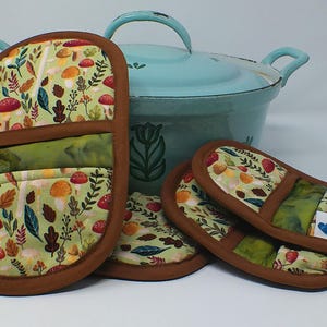 May include: A set of four oven mitts and a teal pot with a lid. The mitts feature a light green fabric with a pattern of mushrooms, leaves, and berries, trimmed with brown fabric. The pot has a handle and a floral design.