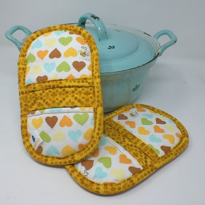 Könnte beinhalten: Zwei gelbe und weiße Topflappen mit einem Herzmuster und einem Bienendesign. Die Topflappen sind aus Stoff und haben eine gesteppte Textur.