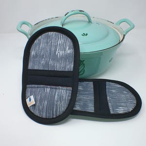May include: Two gray and black oven mitts with a white, vertical stripe pattern. The mitts are oval-shaped with a black border and a small label. A light blue enamel pot with a lid and handles is in the background.