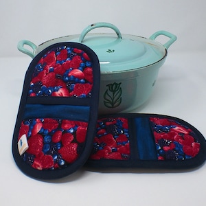Könnte beinhalten: Zwei Topflappen mit Beerenmuster in Blau und Rot. Ein Topflappen ist zusammengefaltet und der andere liegt flach. Im Hintergrund befindet sich ein hellblauer Emailletopf mit grünem Blumendesign.