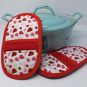Könnte beinhalten: Zwei rot-weiß bedruckte Ofenhandschuhe mit Erdbeeren und ein passender Topflappen. Die Handschuhe und der Topflappen haben rote Verzierungen und Akzente. Im Hintergrund ein hellblauer Topf mit Deckel und Griffen.