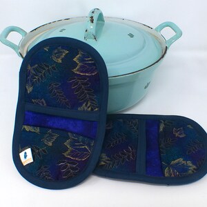 Puede incluir: Dos manoplas de horno azul marino con un estampado de hojas doradas y turquesas, y un ribete azul a juego. Una olla de esmalte azul claro con asas está en el fondo. Las manoplas tienen una banda azul en el centro.