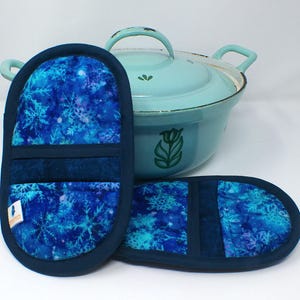 Könnte beinhalten: Zwei blaue Topflappen mit Schneeflockenmuster und ein passender Topf. Die Topflappen haben eine dunkelblaue Umrandung und eine Schlaufe zum Aufhängen. Der Topf ist hellblau mit einem grünen Blumendesign.