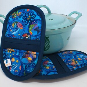 Könnte beinhalten: Zwei marineblaue Ofenhandschuhe mit einem lebendigen blauen, aquatischen Muster mit bunten Fischen und Meeresbewohnern. Im Hintergrund ein hellblauer Topf mit Griffen. Die Handschuhe haben ein Etikett mit einem Logo.