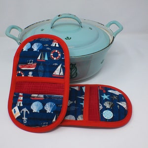 Könnte beinhalten: Zwei blaue und rote Topflappen mit nautischem Thema. Die Topflappen haben einen roten Rand, ein rotes Mittelteil und einen blauen Stoff mit weißen Segelboot-, Leuchtfeuer- und Muschelmotiven.