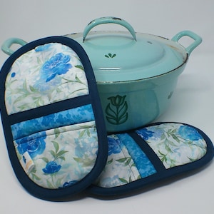 Könnte beinhalten: Ein hellblaues Kochgeschirr mit passendem Deckel und zwei Topflappen. Die Topflappen weisen ein Blumenmuster mit blauen Blumen und grünen Blättern auf weißem Grund auf, das mit marineblauem Stoff eingefasst ist. Das Kochgeschirr hat ein Blumenmuster an der Seite.