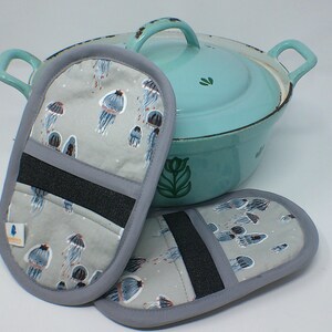 Puede incluir: Una olla esmaltada azul claro con asas y tapa a juego, junto a dos manoplas grises. Las manoplas presentan un estampado de medusas azules y grises. Una banda negra está cosida a través de las manoplas para el agarre.