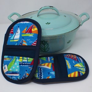 May include: Two blue and white pot holders with a sailboat pattern. The pot holders have a navy blue trim and a pocket on the back. The pot holders are sitting next to a light blue enamel pot with a white floral design.