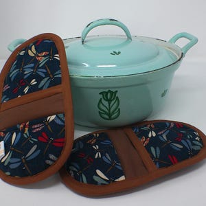 Peut inclure: Une cocotte en fonte émaillée bleu clair avec couvercle, poignées et motif floral. Deux maniques assorties avec un motif de libellules bleu marine, rouge et blanc, et une bordure marron sont au premier plan. Articles de cuisine.