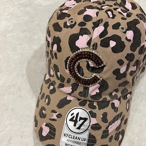 May include: Tan baseball cap with a leopard print pattern in black and pink. The letter "C" is embellished with brown rhinestones. The tag reads "47 Clean Up Adjustable Women's".