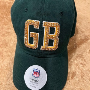 May include: Green baseball cap with a gold "GB" logo adorned with rhinestones. The cap has a white "NFL Team Headwear One Size" label.