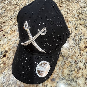 May include: Black baseball cap with silver glitter and a silver rhinestone design of crossed swords. The cap has a black adjustable strap and a New Era logo.