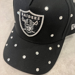 May include: Black baseball cap with a Raiders logo embellished with rhinestones. The cap features a curved brim and is adorned with scattered rhinestones. The Raiders logo is in silver and black. A stylish accessory for sports fans.