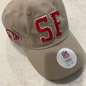 May include: A tan baseball cap with the letters "SF" in red rhinestones. The cap features the San Francisco 49ers logo on the side and an NFL Team Headwear sticker. The hat is one size.