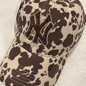 May include: A baseball cap with a brown and tan cow print pattern. The front of the cap features a jeweled logo. The cap has a curved brim and a button on top.