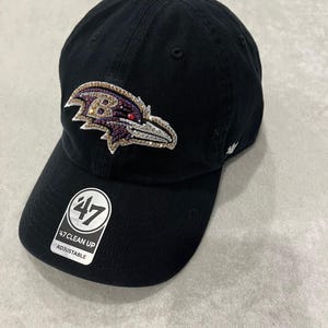 May include: Black baseball cap with an embroidered Baltimore Ravens logo featuring rhinestones. The logo is a purple and gold raven with a red eye. The cap has a 47 Clean Up adjustable strap.