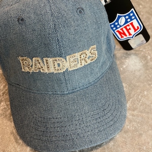 May include: A blue denim baseball cap with a gold sequined "RAIDERS" logo and an NFL tag attached.