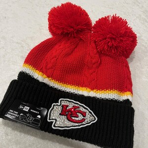May include: Red and black knit beanie with a large rhinestone Kansas City Chiefs logo. The beanie has two red pom poms on top and a white and yellow stripe.