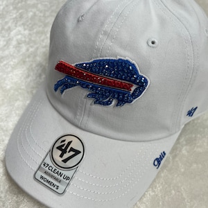 May include: White adjustable baseball cap featuring a blue and red crystal-embellished Buffalo Bills logo. The cap has a curved brim and the word "Bills" embroidered on the side. A tag reads "47 Clean Up Adjustable Women's".