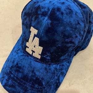 May include: A blue velvet baseball cap with the letters "LA" in silver rhinestones. The cap has a curved brim and a small button on top. The fabric has a textured, crushed velvet appearance.