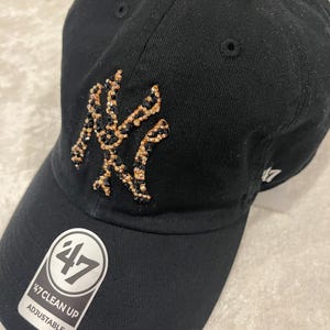 May include: Black adjustable baseball cap with a jeweled New York Yankees logo. The cap features a curved brim and a small logo on the side. The tag reads "47 Clean Up Adjustable".