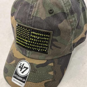 May include: A camouflage baseball cap featuring a rectangular patch on the front adorned with black and yellow rhinestones arranged in a flag pattern. The brim displays a circular sticker with the text 47 CLEAN UP ADJUSTABLE in white and black.