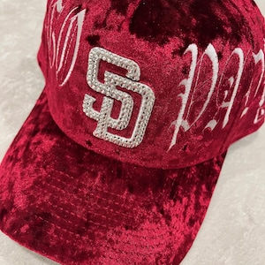 May include: A burgundy velvet baseball cap with the letters "SD" encrusted with rhinestones. The cap also features white embroidered lettering. The hat has a curved brim and a structured crown.